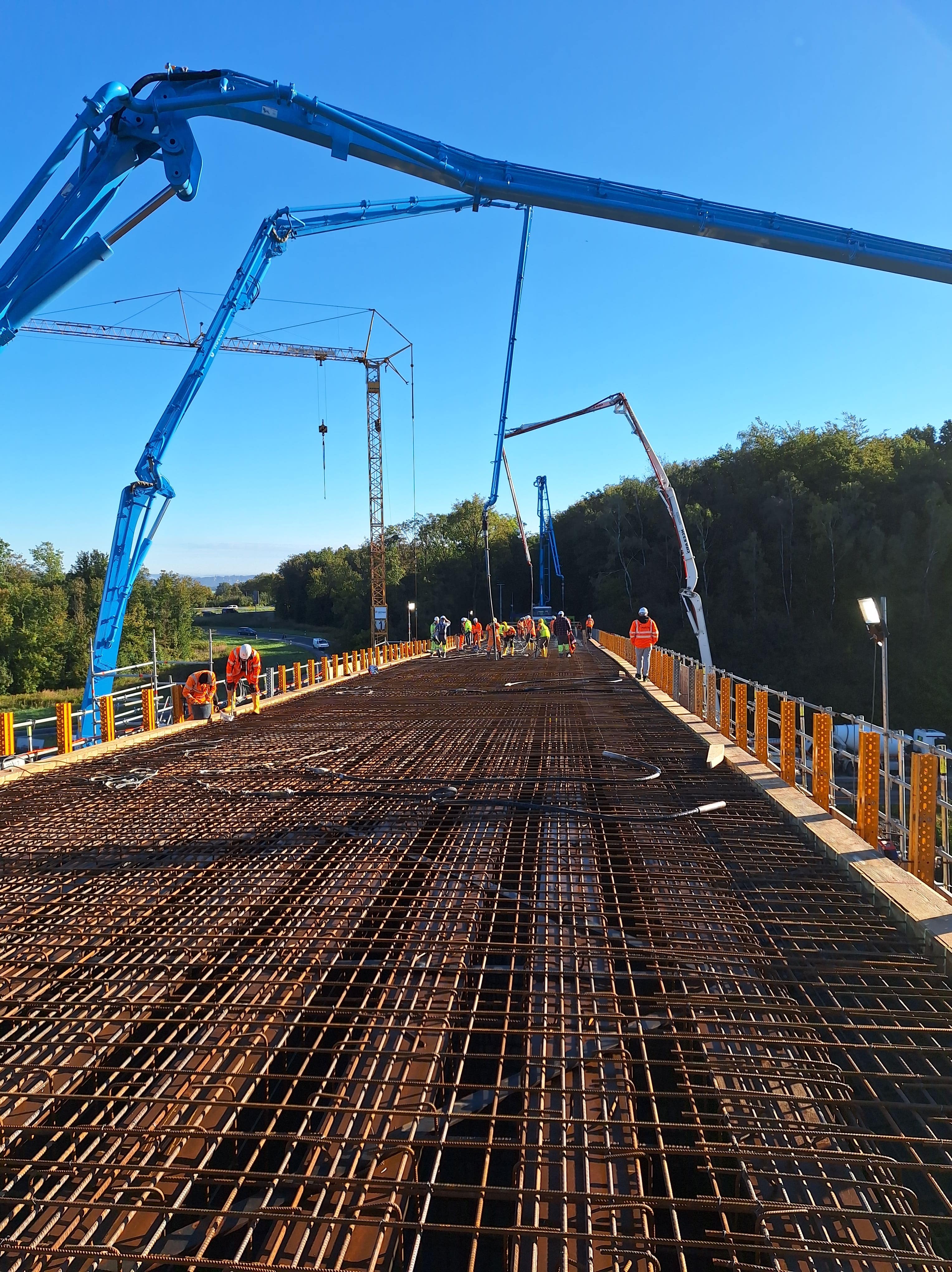 Das Bild zeigt eine Baustelle für den Bau einer Brücke. Im Vordergrund liegt ein enges Stahlgeflecht aus Bewehrungsstäben, das als Grundlage für den Beton dient. Arbeiter in orangenen Warnwesten und Helmen sind auf der Brücke verteilt. Mehrere blaue Betonpumpen mit langen Auslegern ragen in den Himmel. Gelbe Sicherheitsbarrieren säumen die Brücke. Im Hintergrund sind grüne Bäume und ein klarer blauer Himmel zu sehen, was auf eine ländliche Umgebung bei gutem Wetter hindeutet.
                        